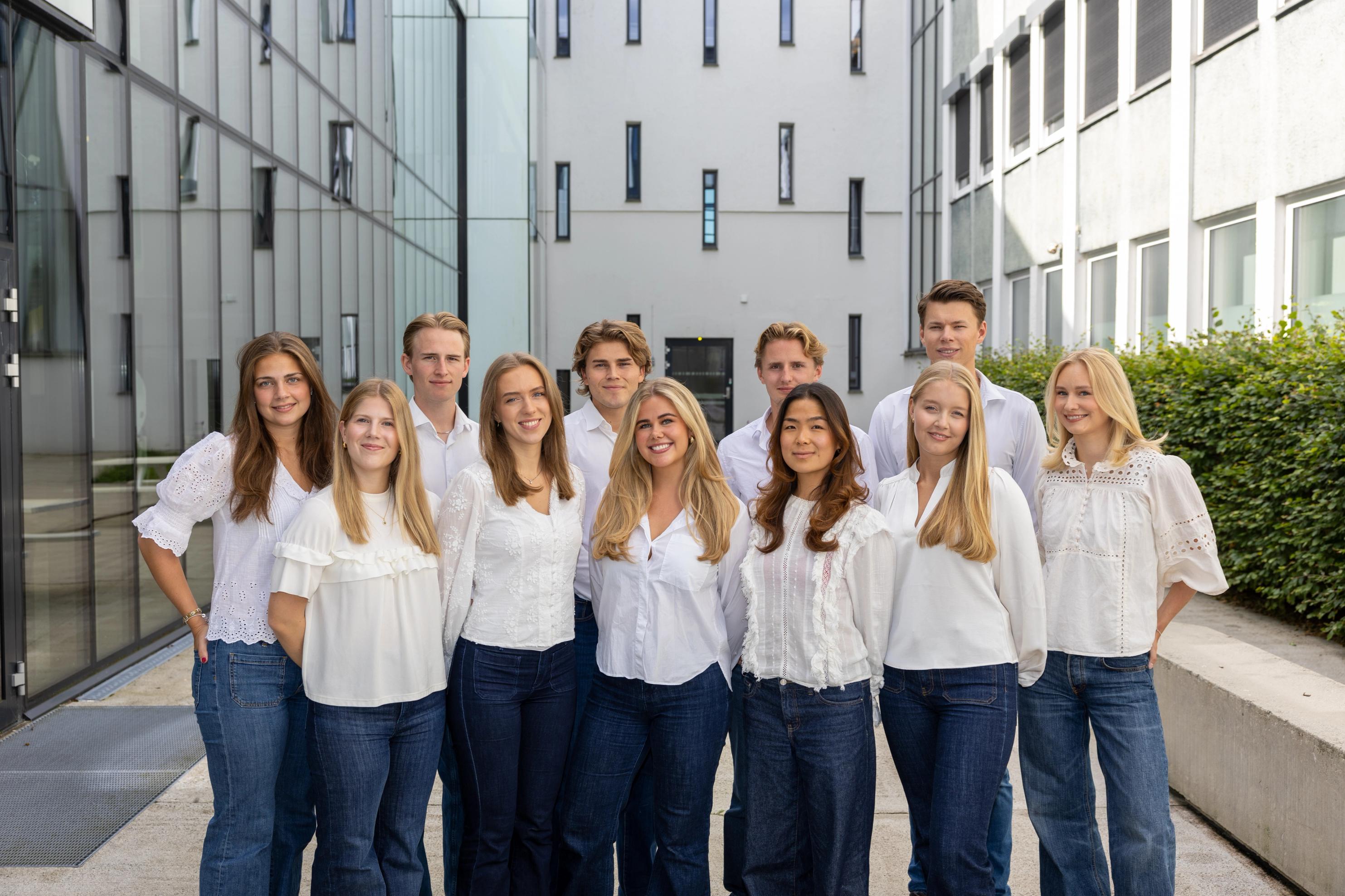 En gruppe unge voksne i hvite skjorter og blå jeans står samlet og smiler foran moderne kontorbygninger.