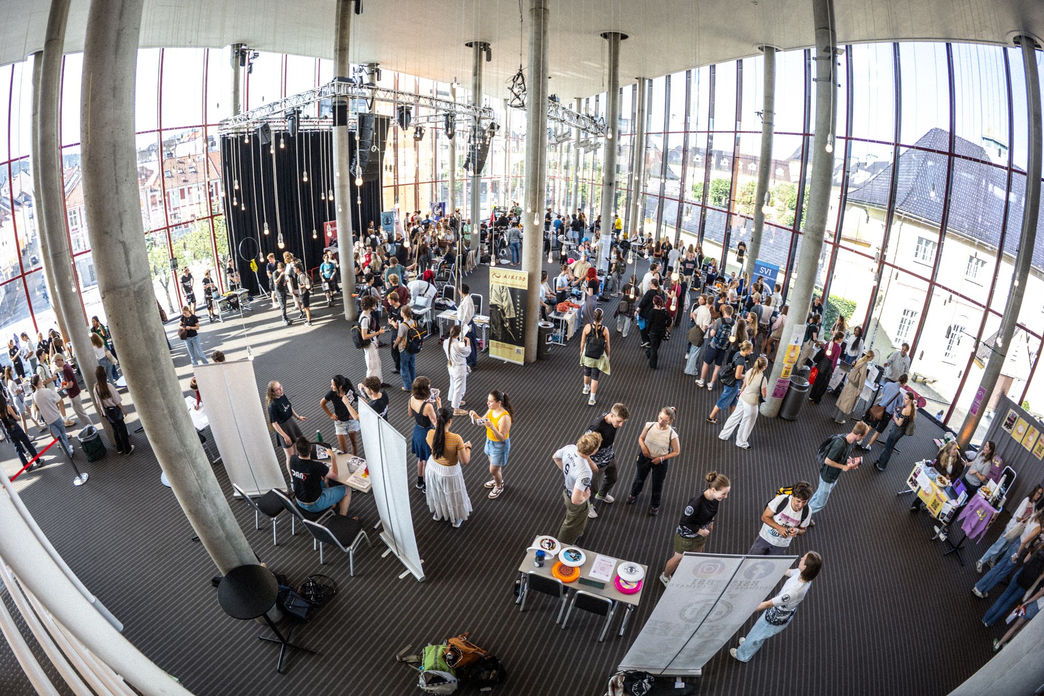 Oversiktsbilde av en lys og moderne messehall der mange studenter går rundt mellom ulike stands og prater med representanter for studentorganisasjoner.