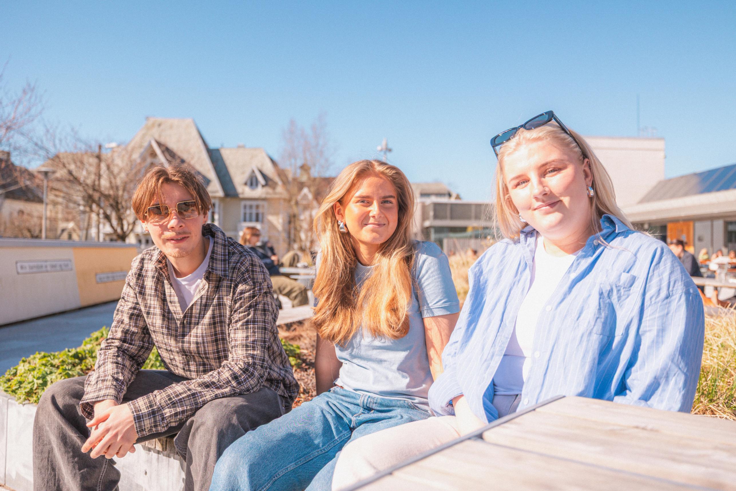 Tre unge personer sitter ute i sola på et campusområde og smiler mot kamera, med moderne bygninger i bakgrunnen.