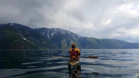 En person padler kajakk på stille vann omgitt av høye, grønne fjell med snøflekker under en overskyet himmel.