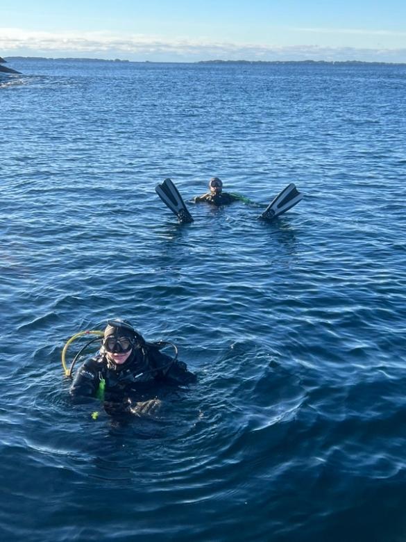 To dykkere flyter i det blå havet under en klar himmel, med svømmeføttene til den bakerste dykkeren synlige over vannflaten.