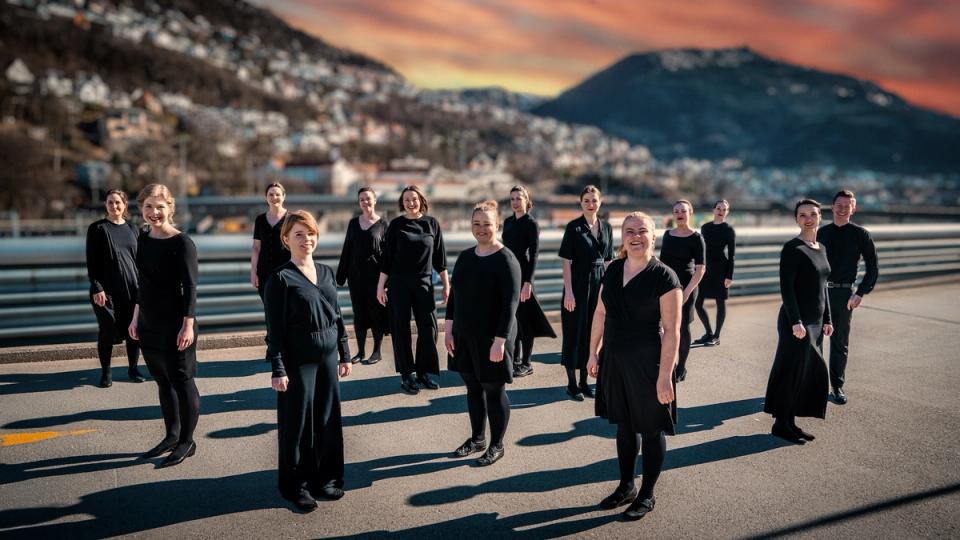 En gruppe mennesker kledd i svarte klær står spredt utover en vei med fjell og bybebyggelse i bakgrunnen under en fargerik solnedgangshimmel.