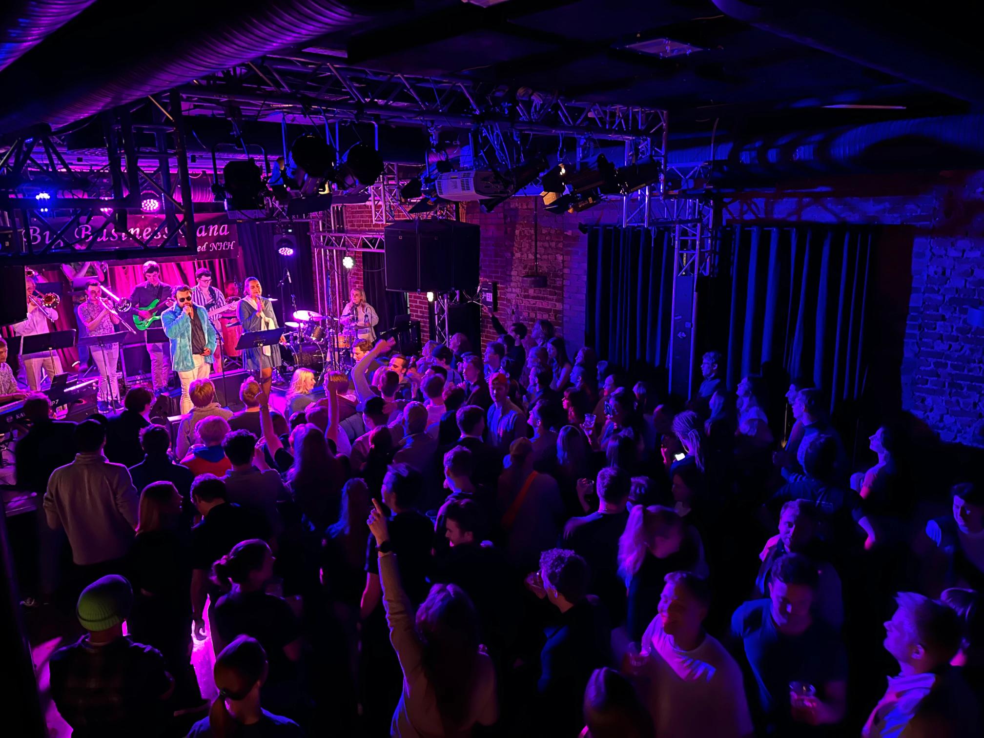 En stor folkemengde stÄr tett sammen pÄ et mÞrkt konsertlokale mens et band spiller pÄ en opplyst scene med fargerike lys. Lokalet har industrielt preg med synlige rÞr i taket og murvegger langs sidene.