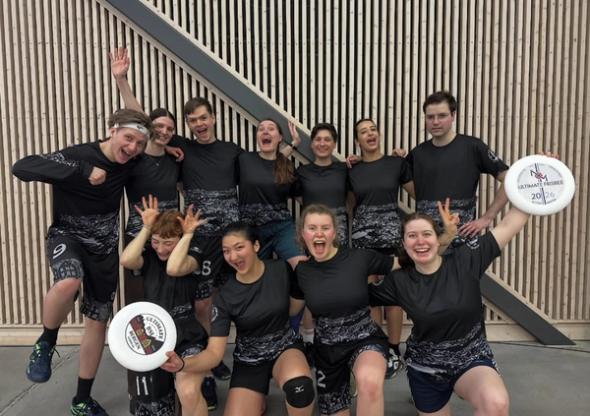 En glad gjeng ultimate frisbee-spillere i matchende svarte drakter poserer og tuller foran en vegg innendørs, mens to av dem holder hver sin frisbee med turneringslogo.