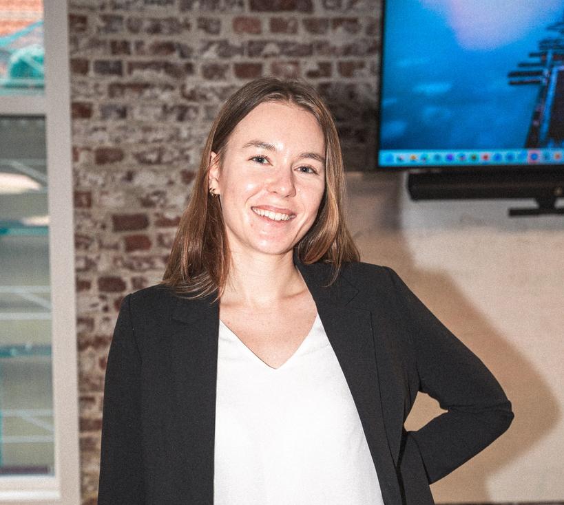 Smilende ung kvinne i hvit bluse og mørk blazer ( Julie) står innendørs foran en murvegg