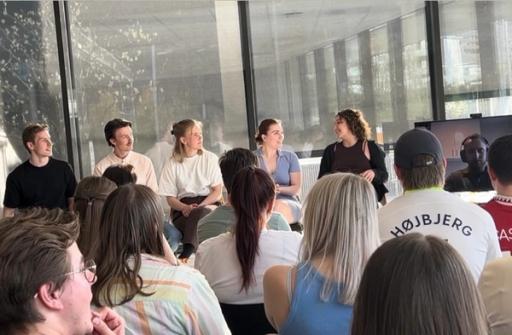 En gruppe unge voksne sitter foran et publikum i et lyst møterom, mens de deltar i en paneldiskusjon eller presentasjon. Publikum sitter tett sammen og følger oppmerksomt med på samtalen.