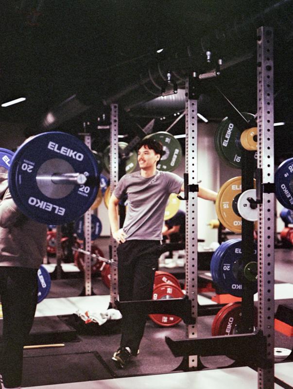 A bright, modern gym with red strength racks, a bench, barbells, and shelves holding colorful kettlebells, dumbbells, and medicine balls.