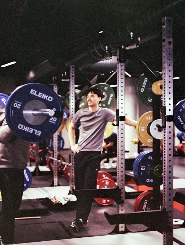 A bright, modern gym with red strength racks, a bench, barbells, and shelves holding colorful kettlebells, dumbbells, and medicine balls.