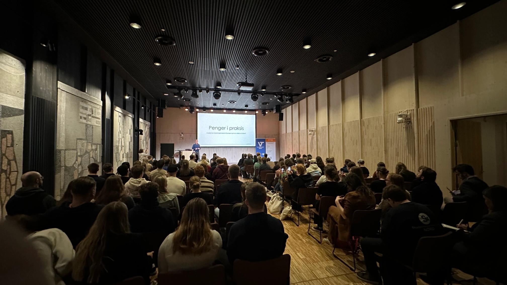 En foredragsholder står foran en stor forsamling i et auditorium og presenterer et lysbilde med teksten «Penger i praksis». Publikum sitter tett i rekker og følger med på presentasjonen i det moderne konferanserommet.