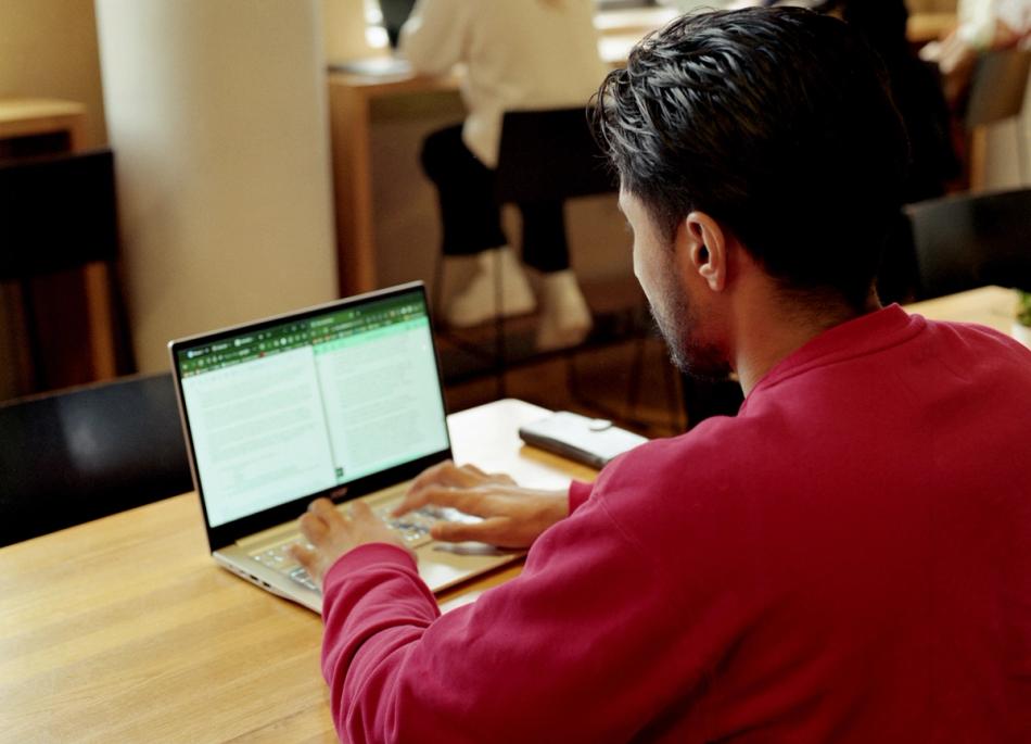 Person som sitter ved et trebord og jobber på en bærbar PC med delt skjerm i et åpent kontor- eller studiemiljø.
