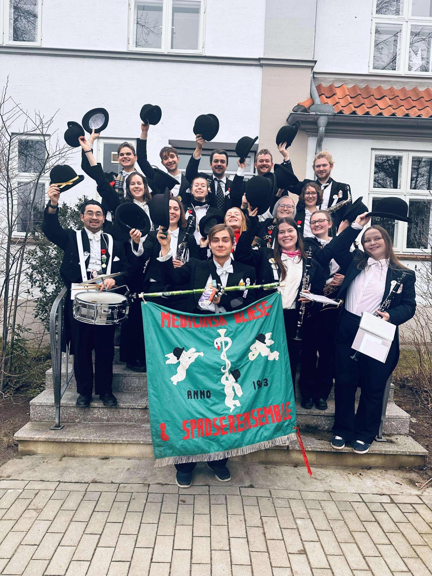 En glad gjeng musikanter i uniform står på en trapp foran en bygning, løfter hattene i været og holder et grønt banner med teksten «Medisinsk Blæse- & Spadserensemble».