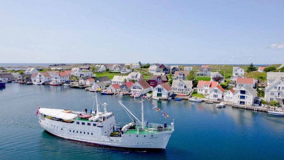 Et hvitt passasjerskip seiler gjennom en rolig havn omgitt av små, fargerike trehus og båter langs kaiene på en solrik dag.