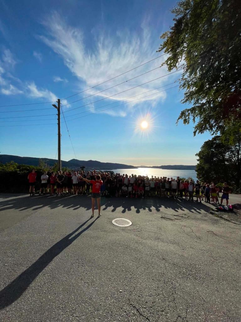 En stor gruppe løpere står samlet på en solrik vei med utsikt over en fjord, mens en person i rød T-skjorte foran dem gir instruksjoner før et løp.