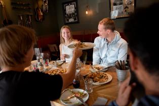 En vennegjeng sitter rundt et bord på en restaurant og deler pizza, mens en person rekker et fat over bordet.