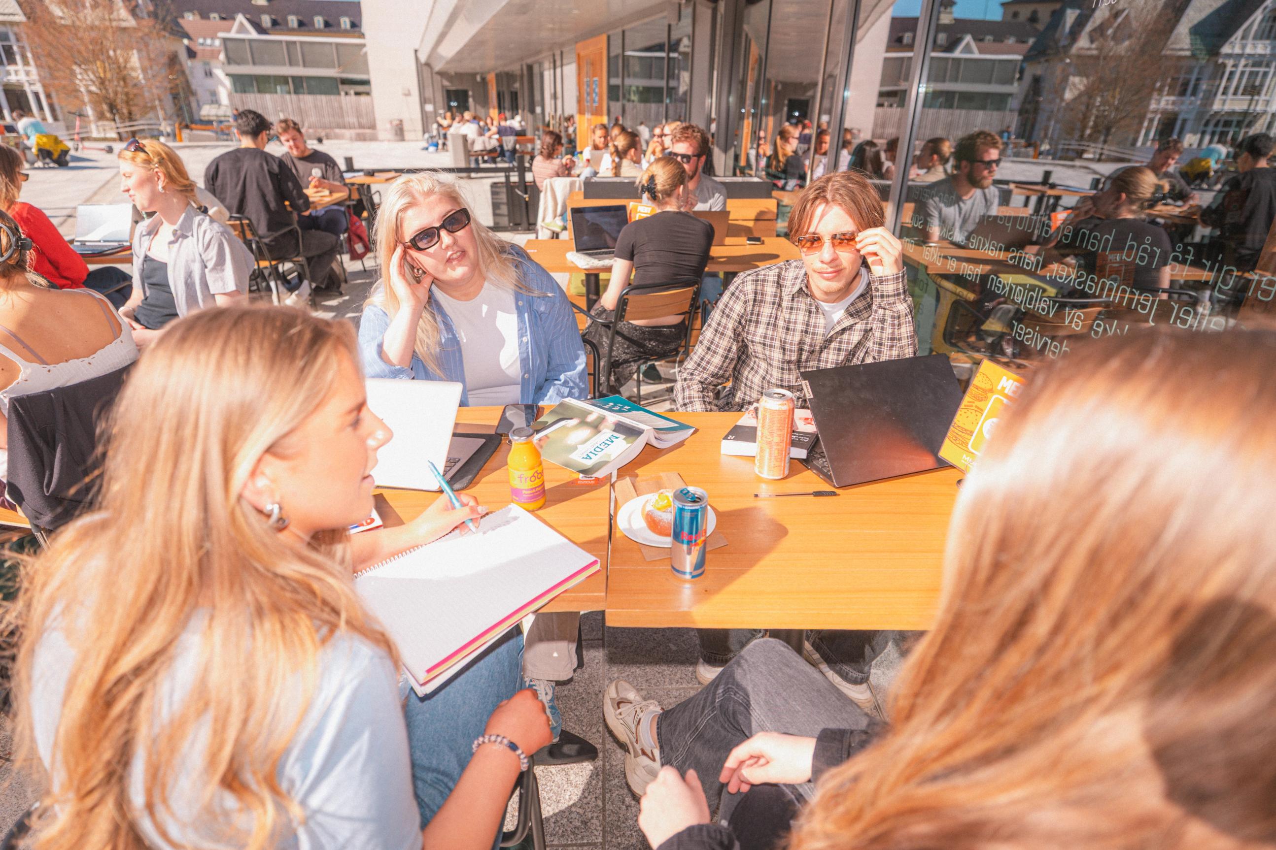 En gruppe studenter sitter ute på en solfylt kafé eller campusområde, der de prater, skriver i notatbøker og jobber på bærbare PC-er.