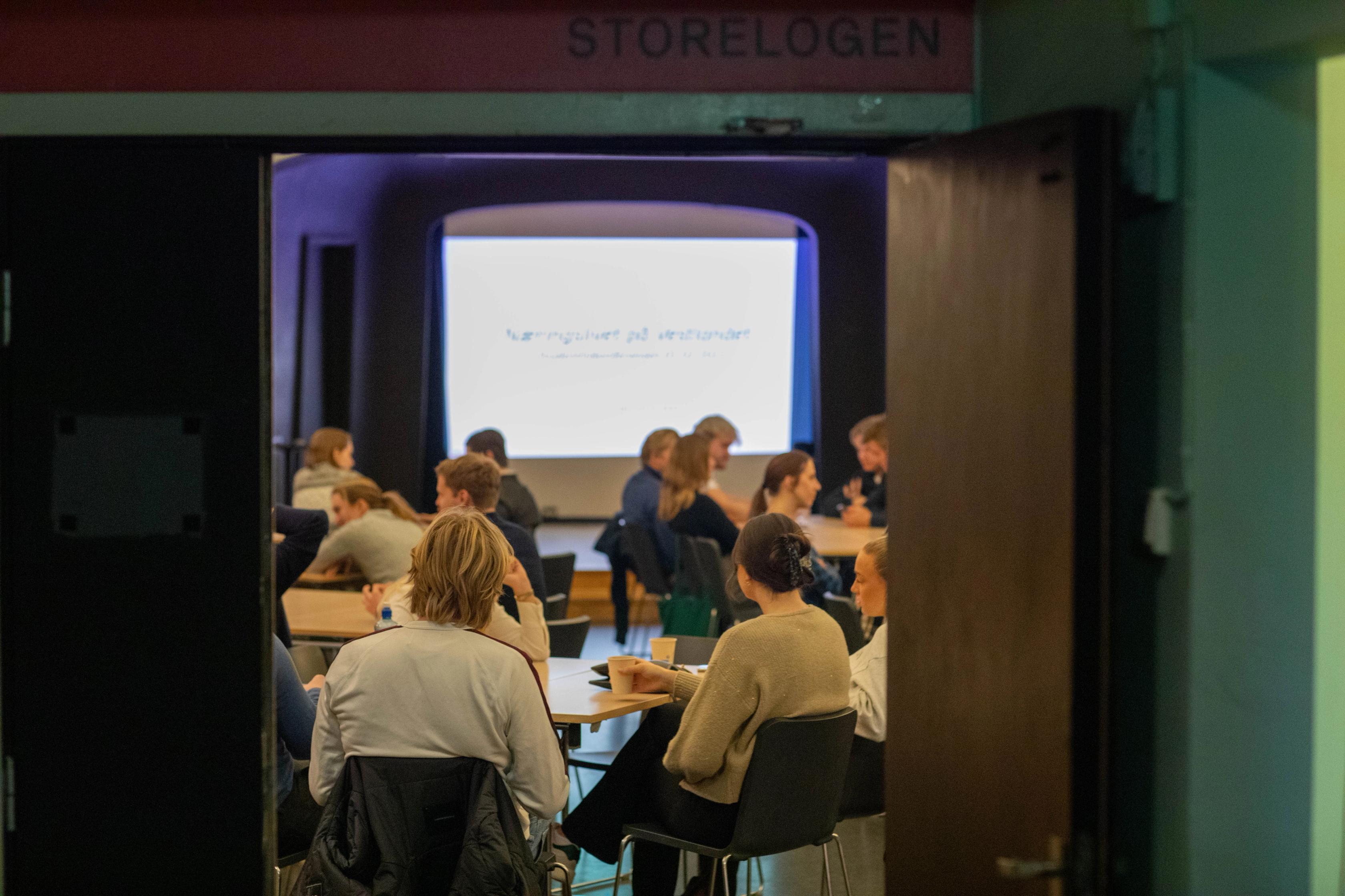 En gruppe mennesker sitter rundt bord i et rom merket «Storelogen» og følger med på en presentasjon som vises på en stor skjerm foran i lokalet.