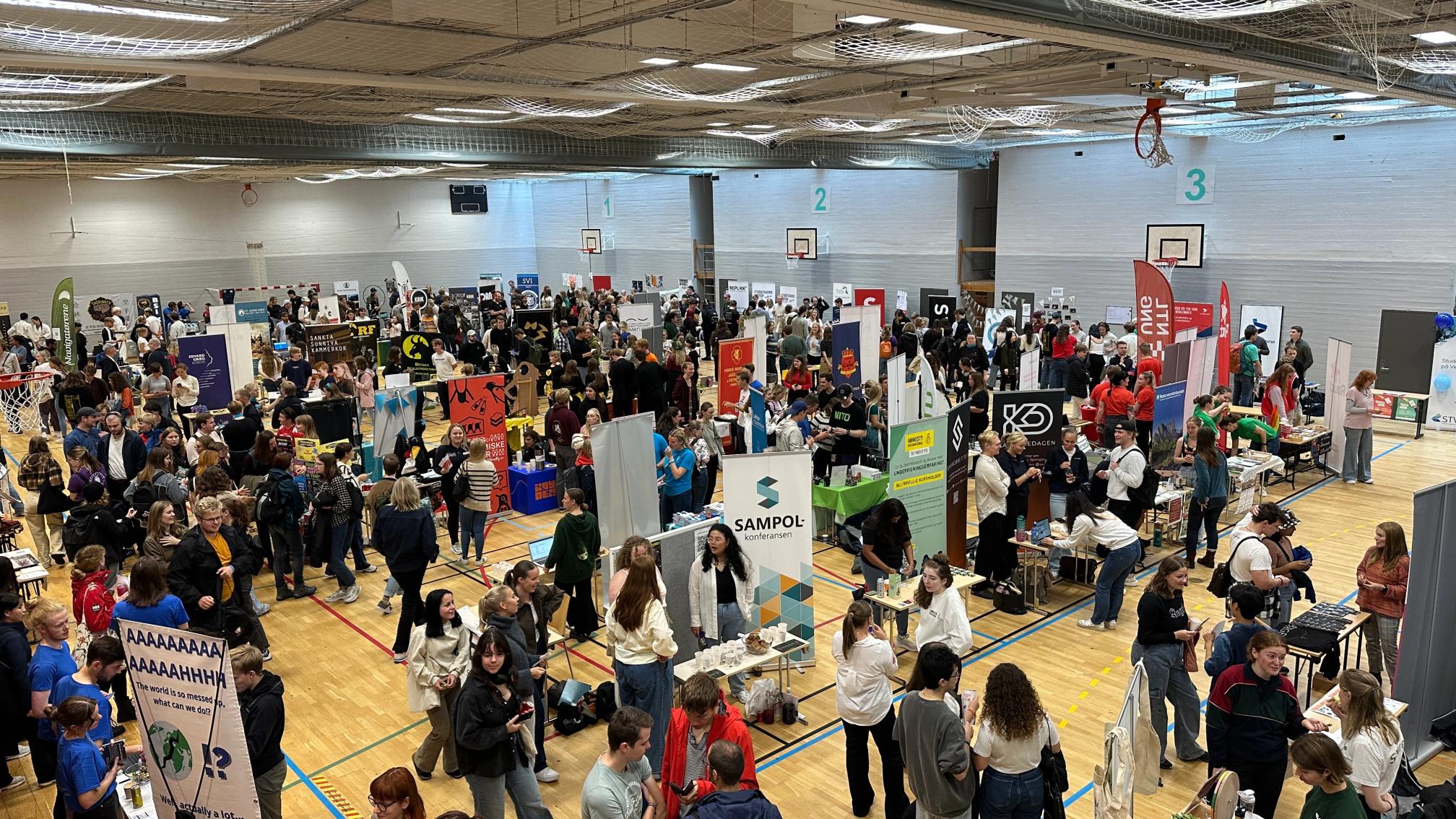 En fullsatt messe i en idrettshall, der mange mennesker besøker ulike stands og snakker sammen.