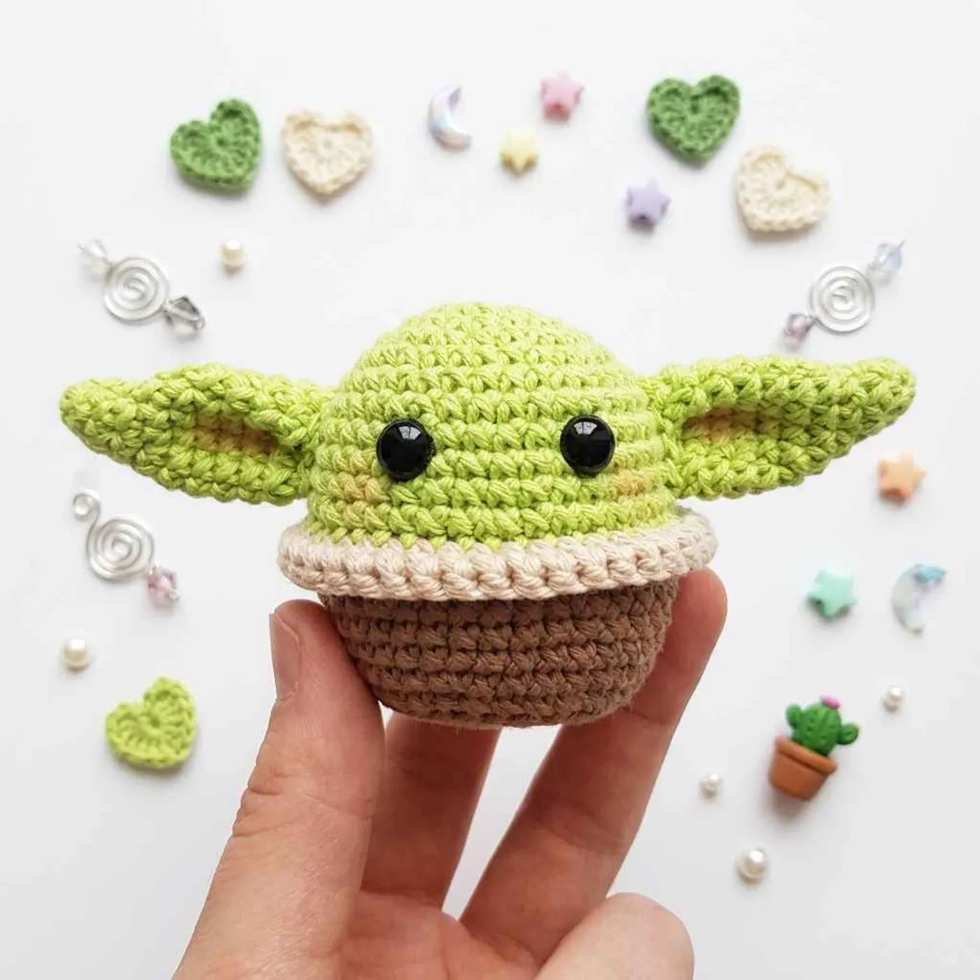 Small green crochet Baby Yoda amigurumi sitting in a brown crochet cup with a beige ruffled rim, with black eyes and pointed ears with a tan inside, held in a hand.