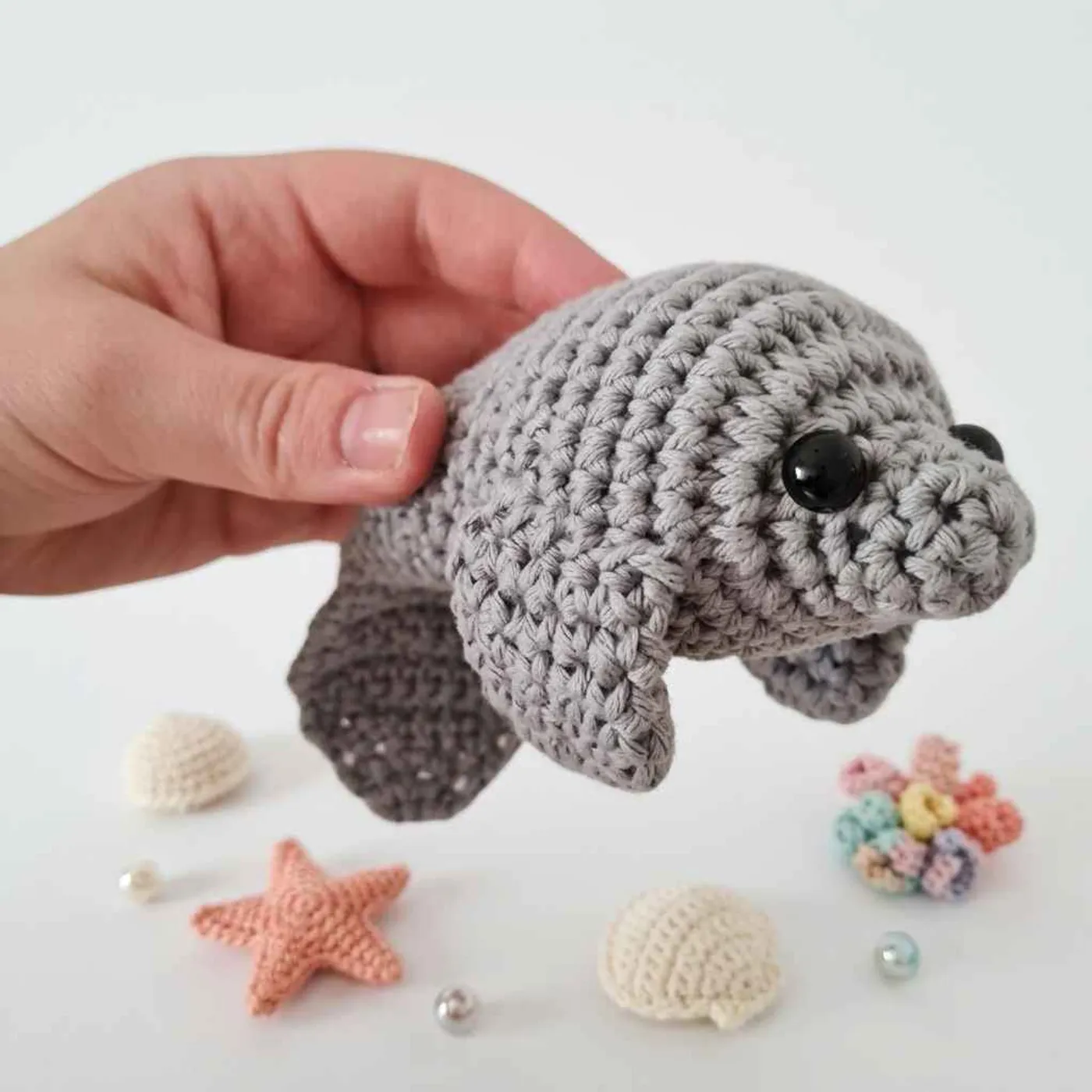 A small grey crocheted manatee with black safety eyes, two grey side fins and a flat rounded tail, held in the palm of a hand