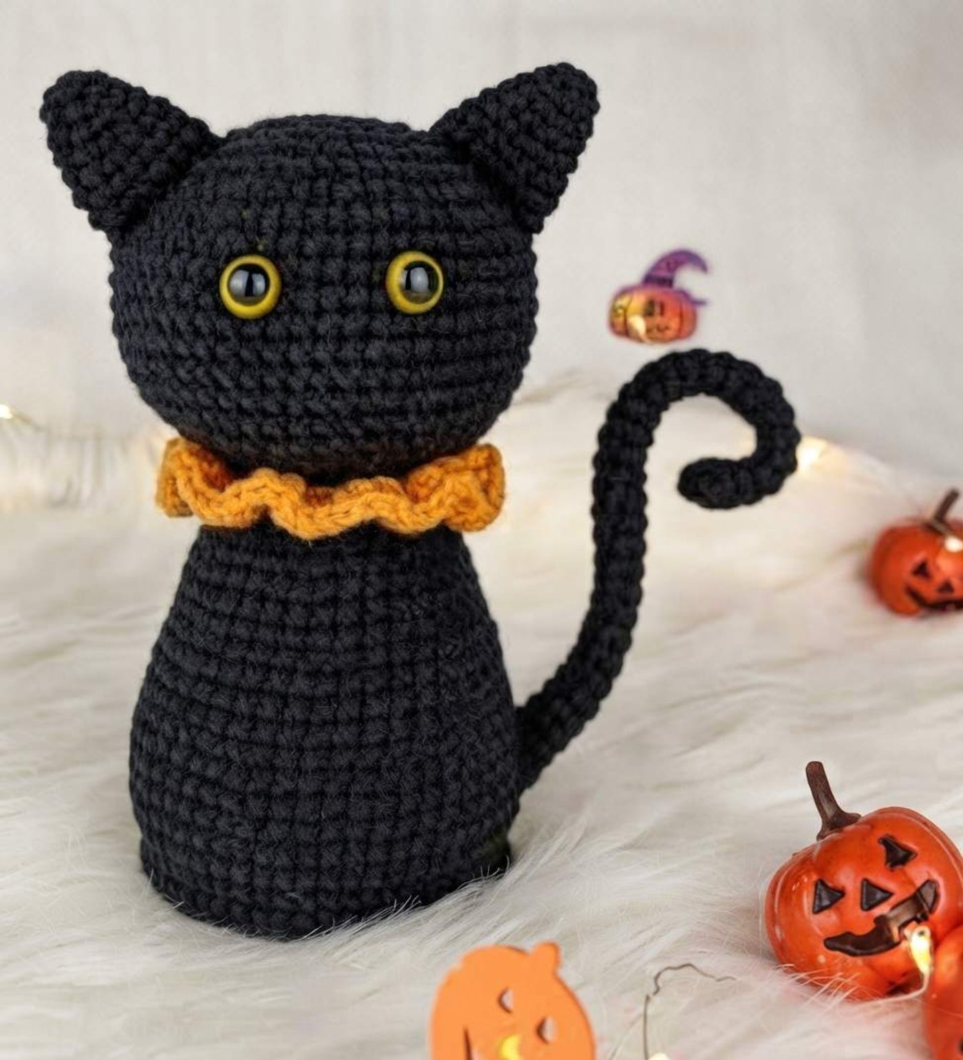 Crocheted black cat amigurumi sitting upright with a round head, two pointed ears, yellow safety eyes, an orange ruffled collar and a long tail curled into a spiral at the tip.