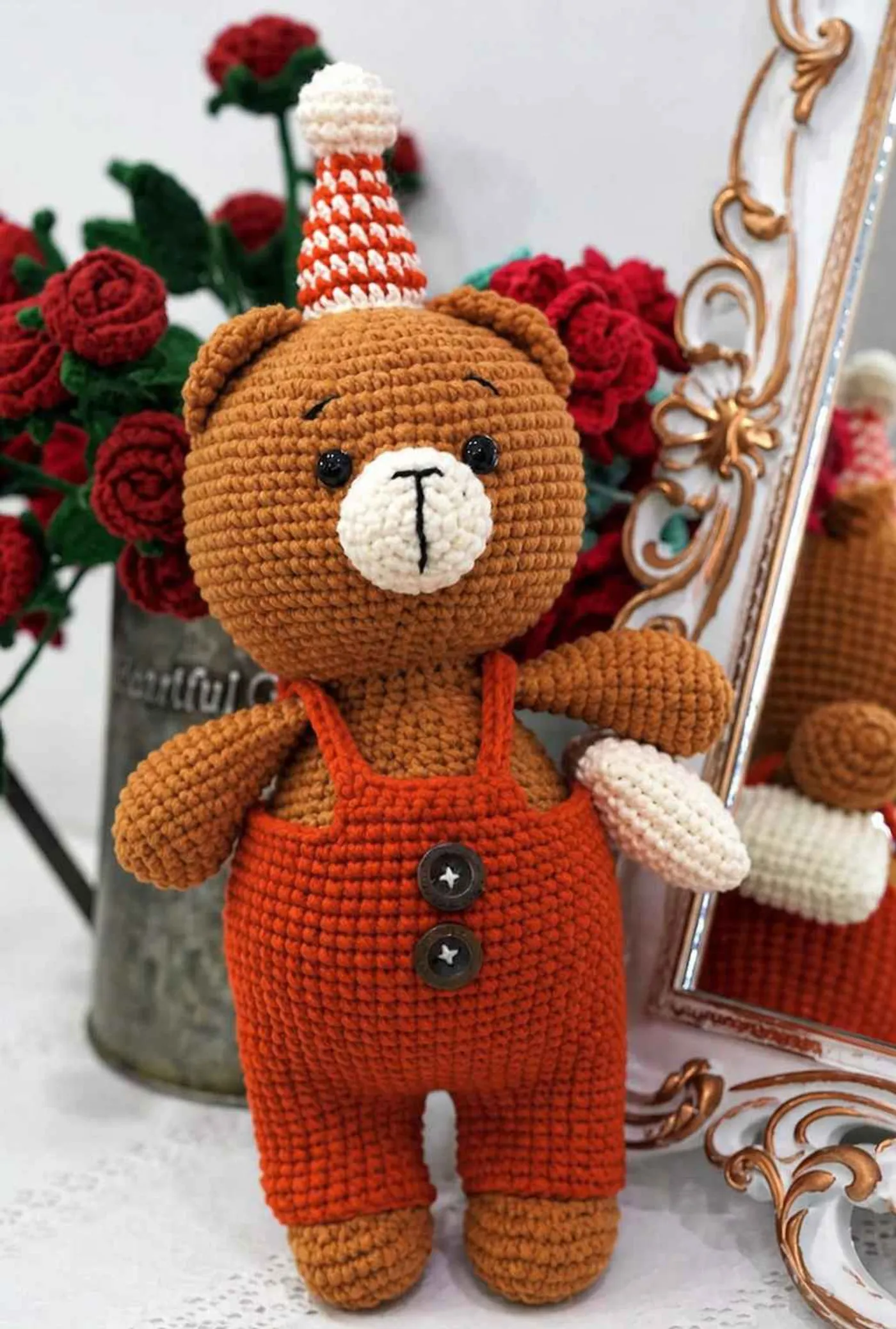 An adorable brown amigurumi crochet bear wearing bright orange overalls and a striped party hat, holding a tiny white pillow - Free pattern.