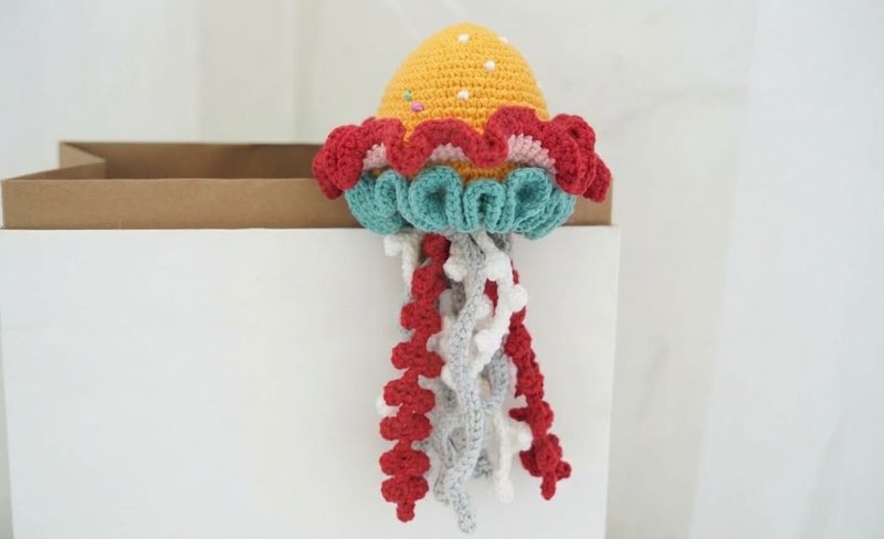 Handmade crochet jellyfish with a golden yellow bell, layered pink, red and teal ruffles, and long red, white and grey tentacles with little curls, resting on the edge of a paper bag.