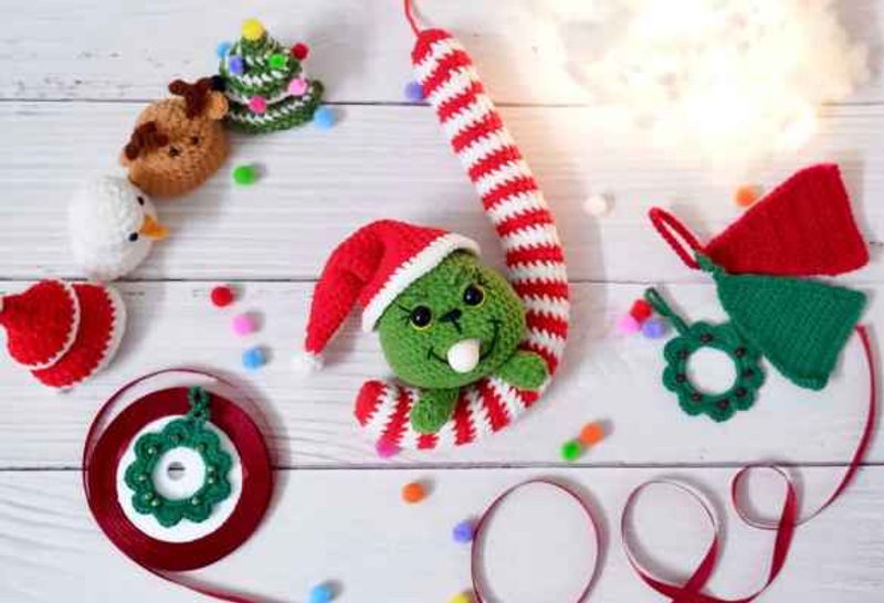 Green crochet Grinch head with black safety eyes, a stitched smile and a white inflatable ball in his mouth, wearing a red Santa hat with a white brim and pompon, beside a long red and white striped c