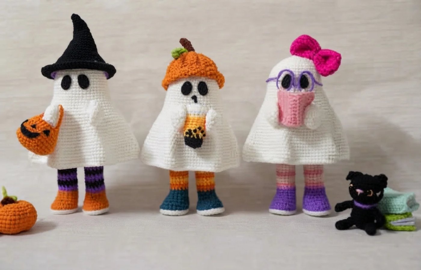 Three crocheted white ghost dolls with striped legs and little boots: one in a black witch hat holding an orange pumpkin basket, one in a pumpkin hat holding a boba milk tea, and one with a pink bow a
