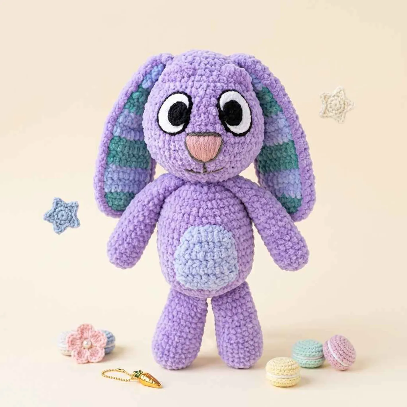 Lilac crochet bunny plush sitting up, with long floppy ears lined in blue and green stripes, large white eyes with black pupils, a pink nose, a light blue belly patch and long arms and legs.