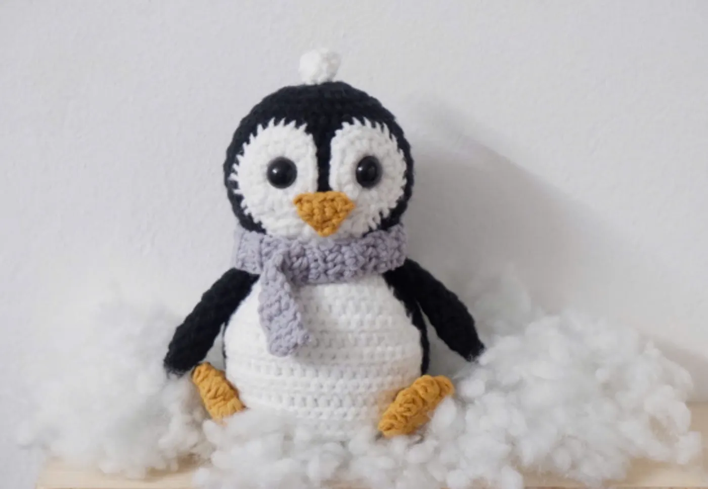 A handmade crochet penguin with a grey scarf, white belly, and yellow feet. Free pattern for beginners.