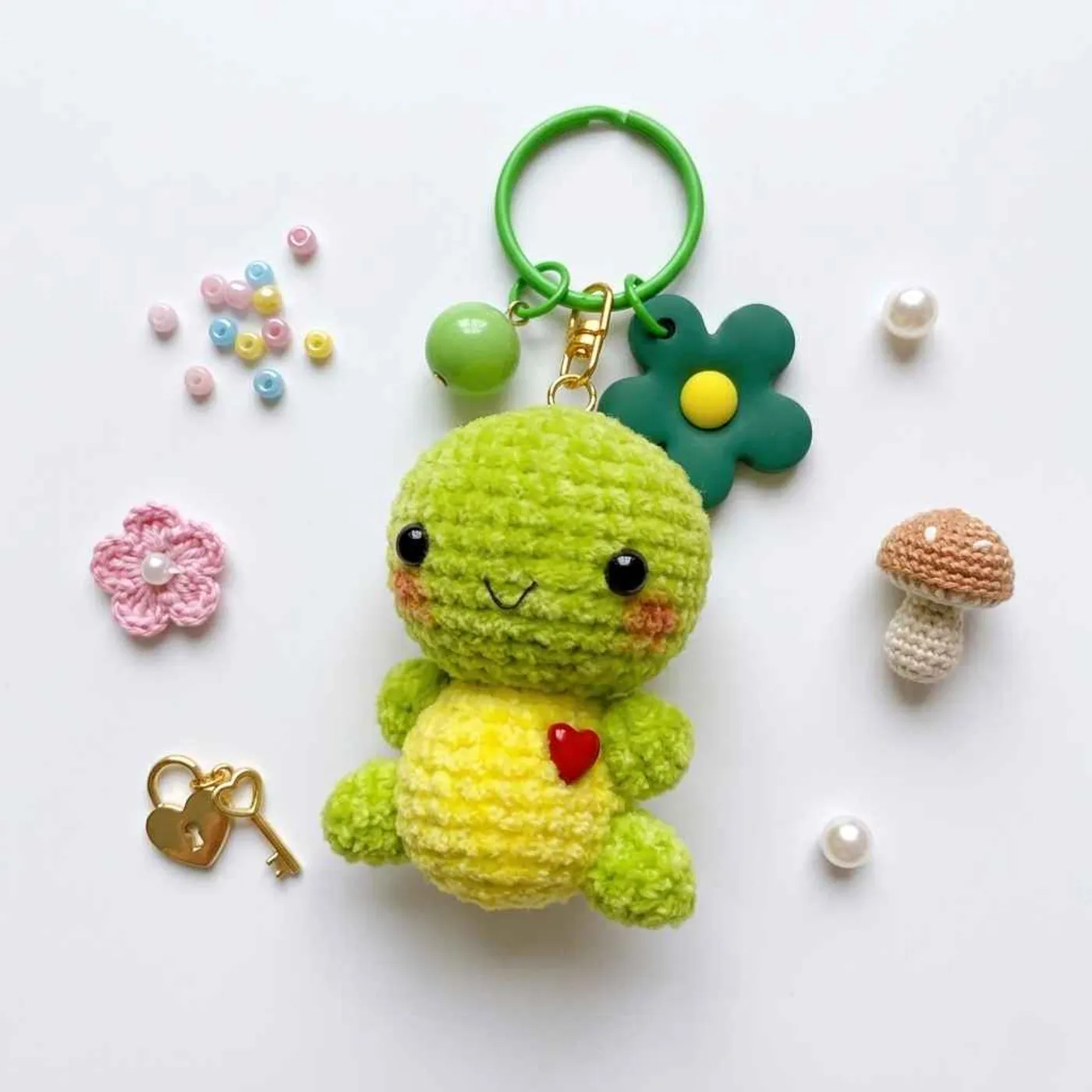 Small green crochet turtle keychain with a round green head, black eyes, a stitched smile and pink cheeks, a yellow belly with a small red heart, and green hands and legs, hanging from a green ring wi