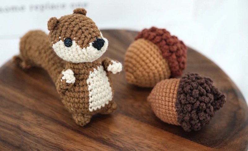 Handmade brown crochet squirrel amigurumi with a cream muzzle and belly, small round ears, black eyes and nose, standing upright with little arms out and a long stuffed tail, next to two crochet acorn