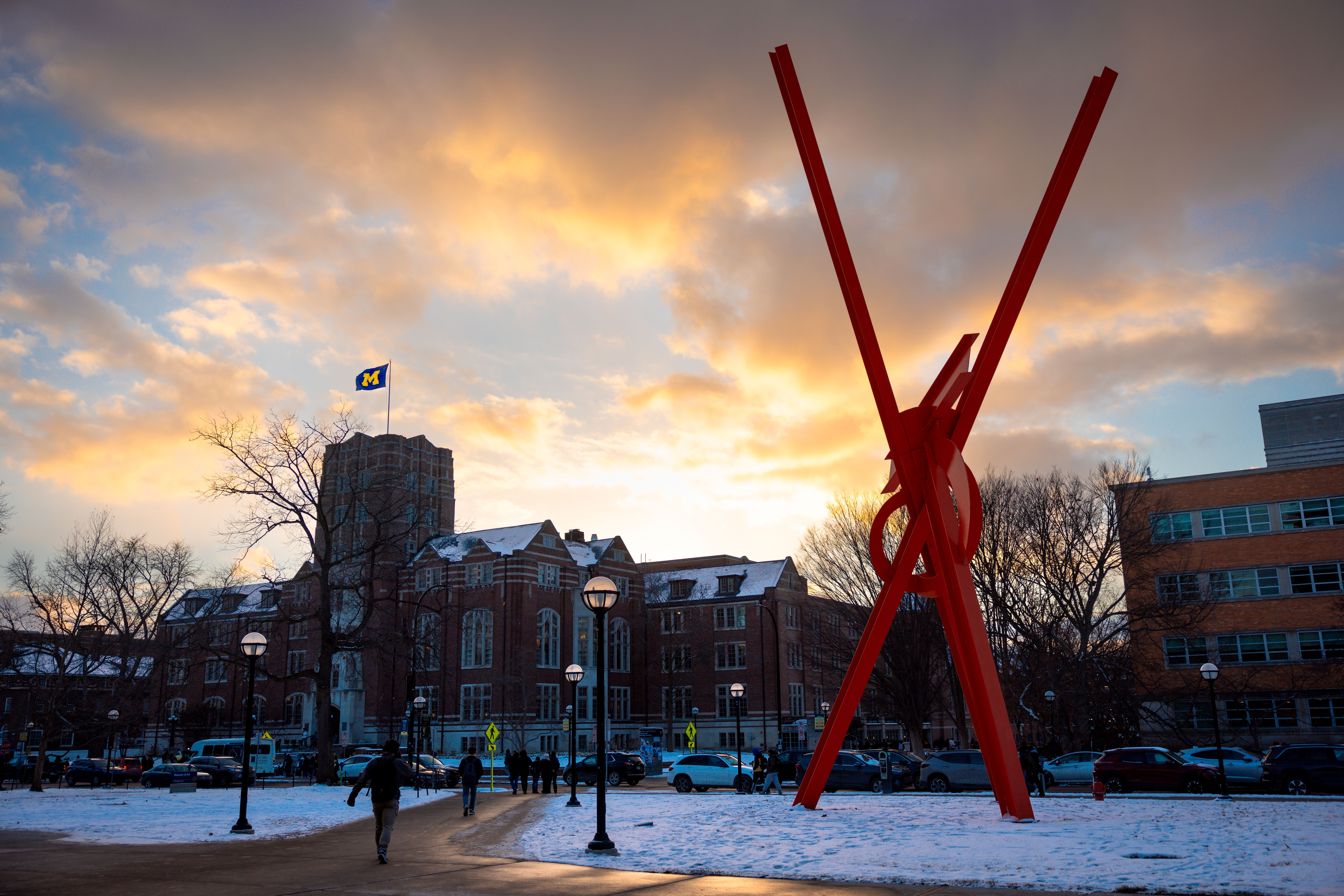 A snowy winter sunset on the University of Michigan campus, with students walking past the red “Begob” sculpture and the Michigan flag flying in the cold evening light