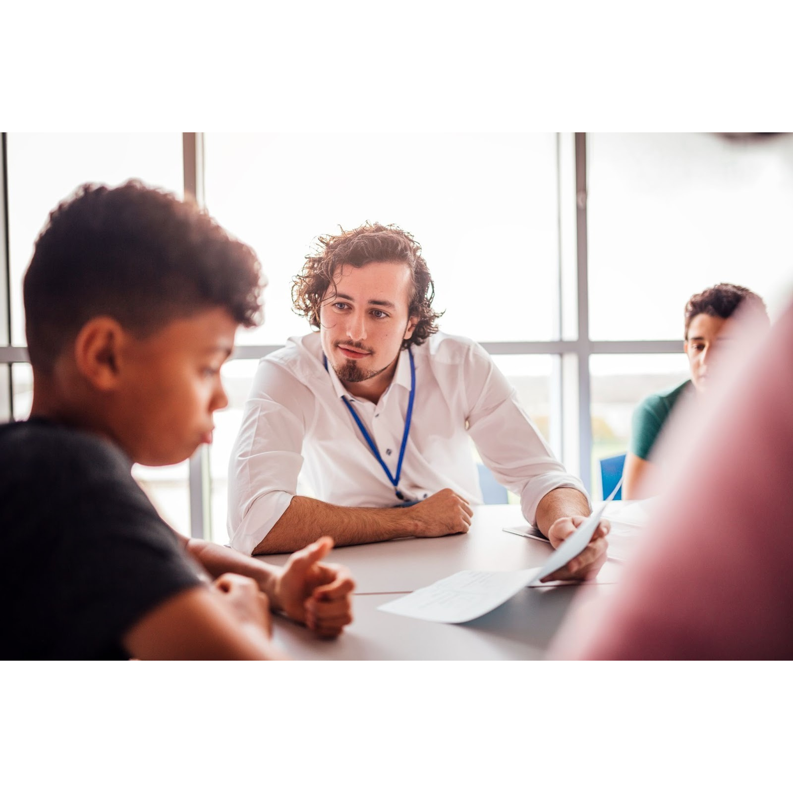 Adult educator or mentor in conversation with two young students at a table in a bright classroom setting, demonstrating engaged listening and supportive dialogue