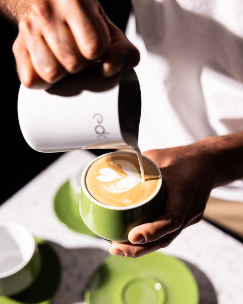 A barista pouring steamed milk into a green cup, drawing a heart in the latte art