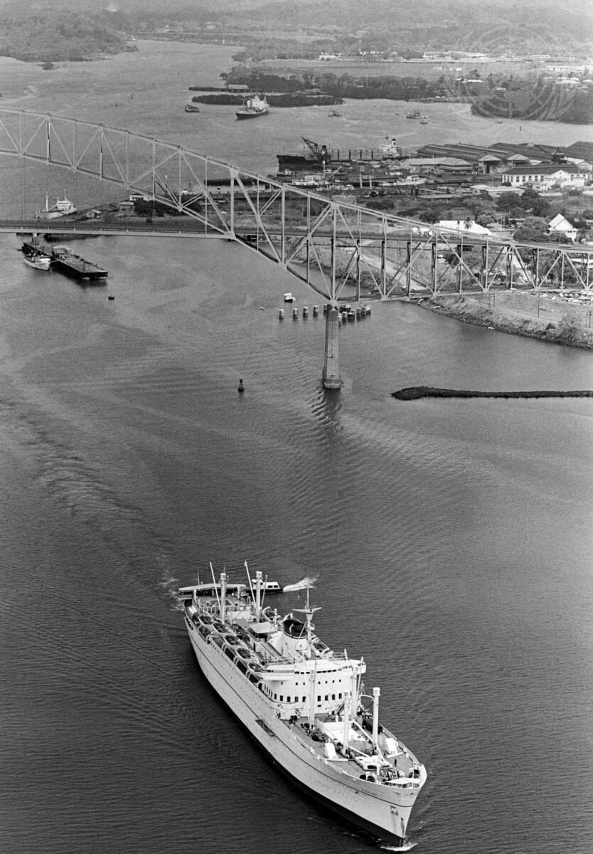 Fotografía en blanco y negro de un buque saliendo de un puerto bajo un puente