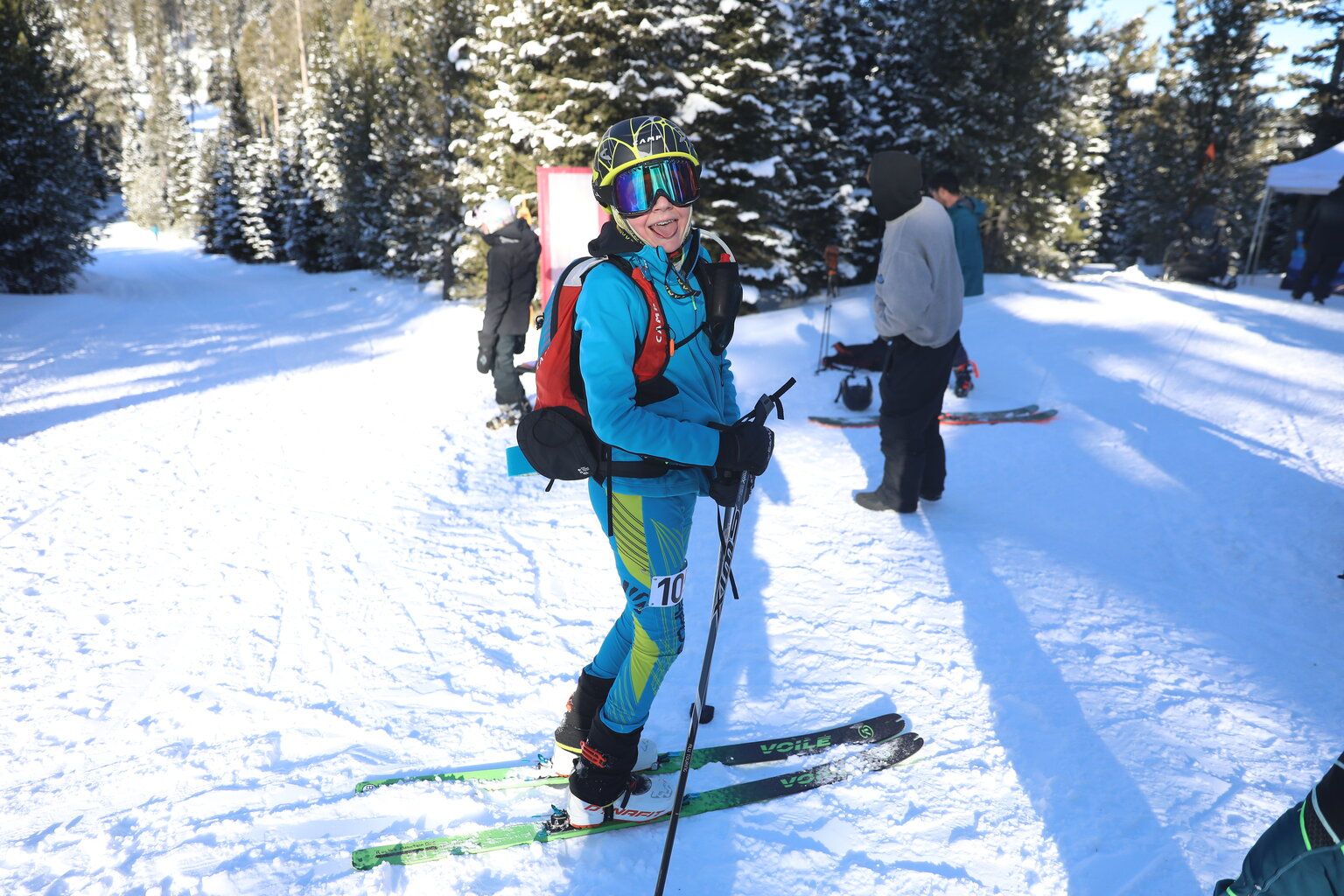 Skier in a blue race suit smiling on a snowy trail with trees and other skiers in the background