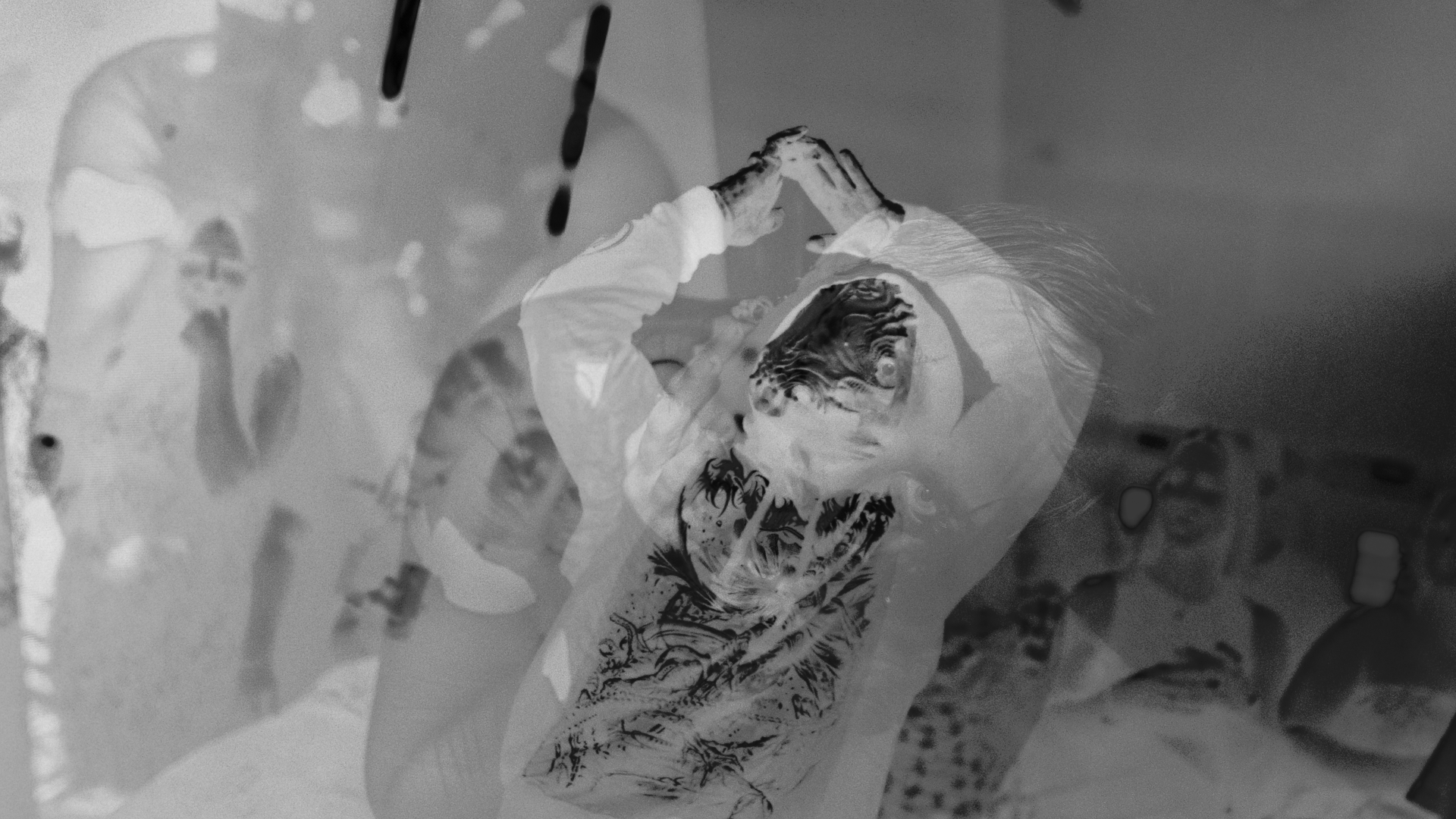 An abstract black-and-white image with an inverted, negative-film effect. A figure raises both arms above their head, hands meeting at the fingertips, their face and body distorted by overlapping ghostly layers and dark ink-like smudges.