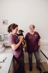 Dr. Kirsten Penner und Anna Friedrichs im Behandlungsraum mit einem Hund auf dem Arm.