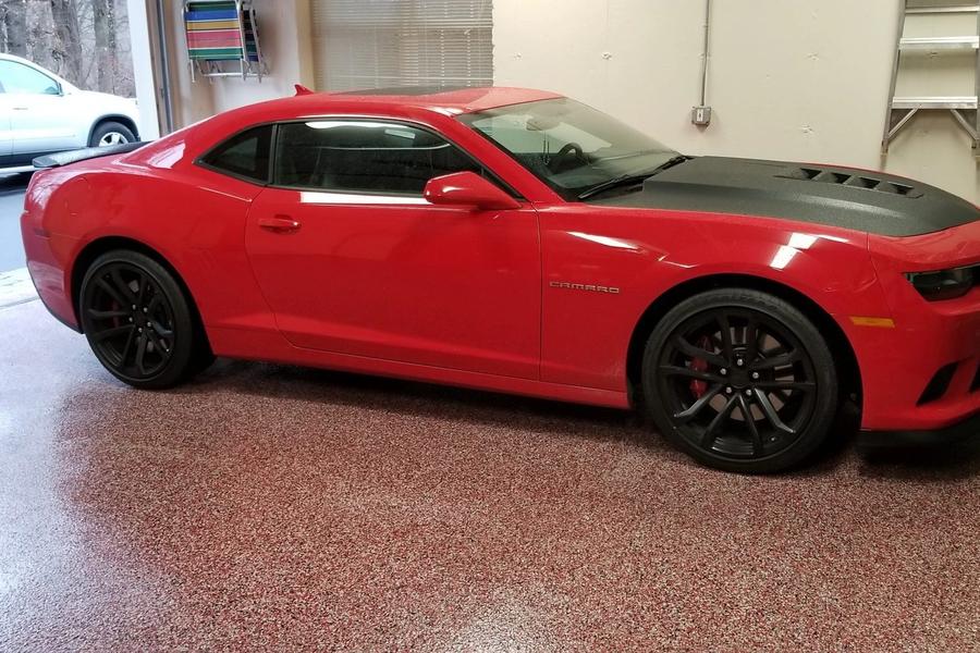 Red Chevrolet Camaro with black wheels parked on a glossy red-flecked epoxy garage floor, with an SUV visible in the driveway outside.