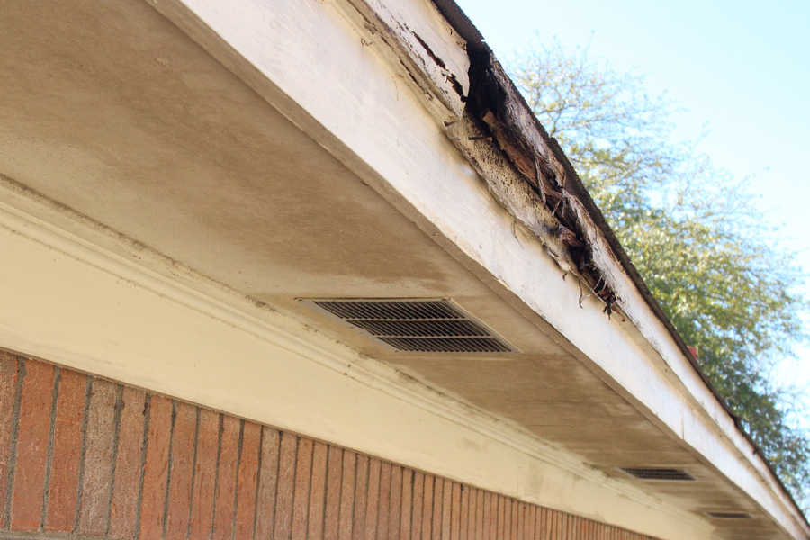 Close-up of a home's cream-colored fascia board and soffit with a section of rotted, crumbling wood along the roofline above a brick wall.