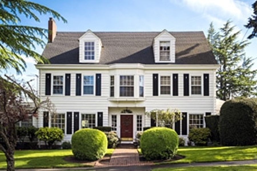 White colonial-style house with black shutters, dormer windows, and a red front door, framed by trimmed round hedges and a brick walkway.