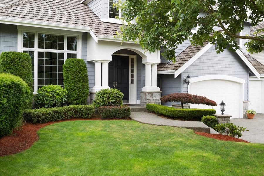 Front entrance of a blue-gray shingled home with white columns, a black front door, white garage door, and trimmed shrubs along a green lawn.