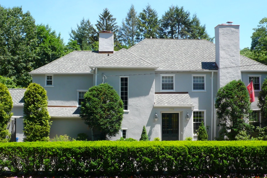 Two-story light gray stucco home with white-trimmed windows, twin chimneys, trimmed hedges, and tall shrubs under a clear blue sky.