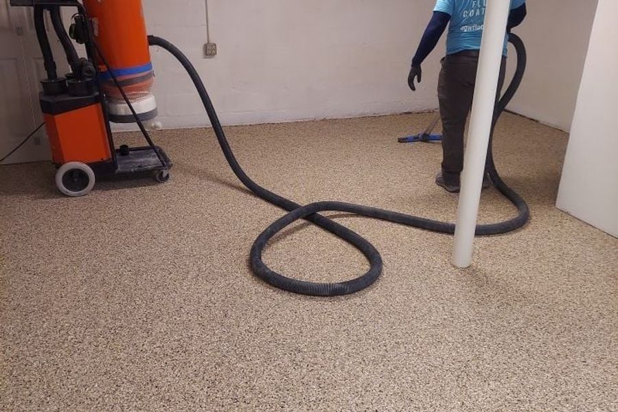 Worker in a blue floor-coatings shirt using a dust-extraction vacuum with a long hose on a newly coated tan flake basement floor with white walls.