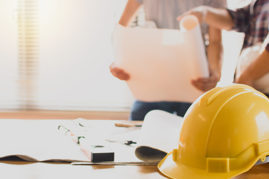 Yellow hard hat resting on a table with rolled blueprints and tools while two people review a large plan sheet in the blurred background.