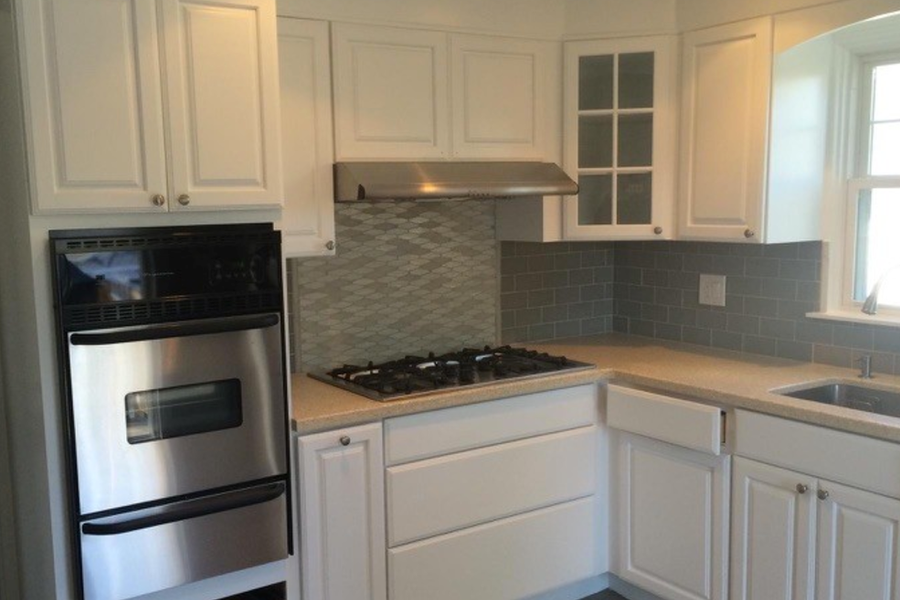 Kitchen with white painted cabinets, a stainless steel wall oven and range hood, gray tile backsplash, and a window over the sink.