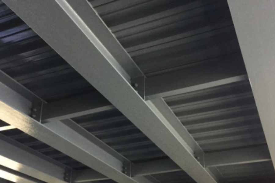 Underside of a structural steel ceiling with gray-painted I-beams supporting corrugated metal decking, viewed from below at an angle.