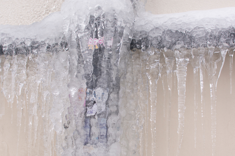 An exterior pipe on a beige wall encased in thick ice, with long icicles hanging from the frozen horizontal line above it.