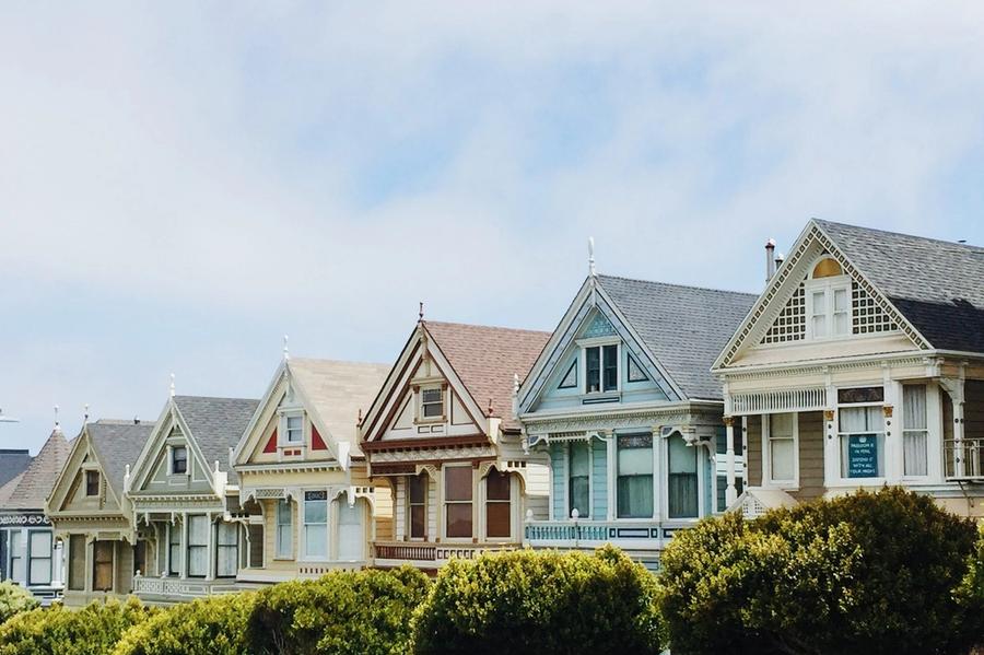 Row of Victorian houses painted in pastel greens, blues, and creams with ornate gabled rooflines, set behind rounded trees under a hazy sky.